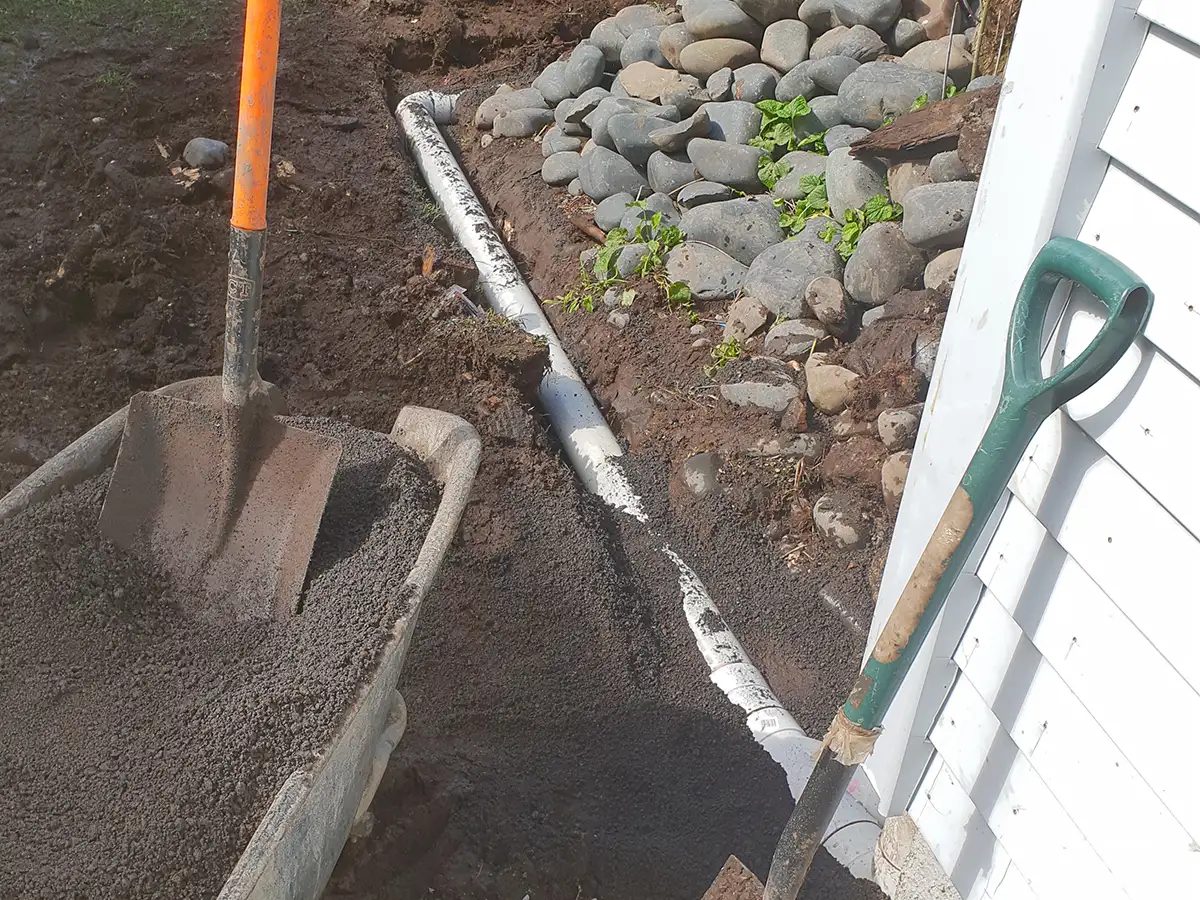 New PVC drain pipe being laid and bedded in a trench beside a house in Whangarei, with a wheelbarrow of bedding metal